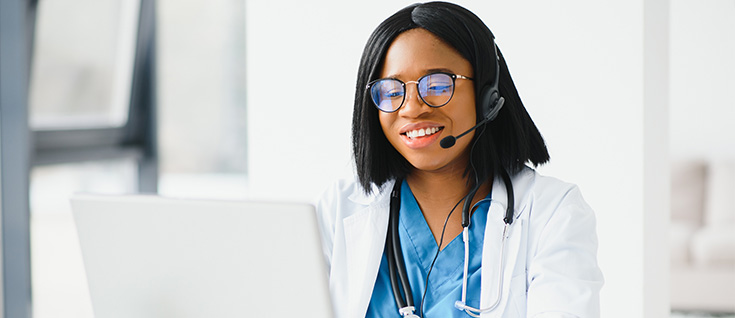 A female doctor smiling while looking at a laptop screen and smiling with glasses and a stethoscope around her neck.