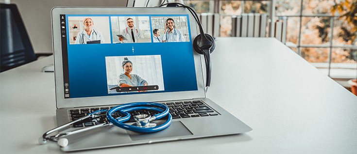 Laptop showing a telemedicine video call with doctors and a patient, with a stethoscope and headset on the desk.